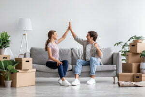 Happy caucasian millennial lady and guy sit on sofa give high five in room interior among boxes